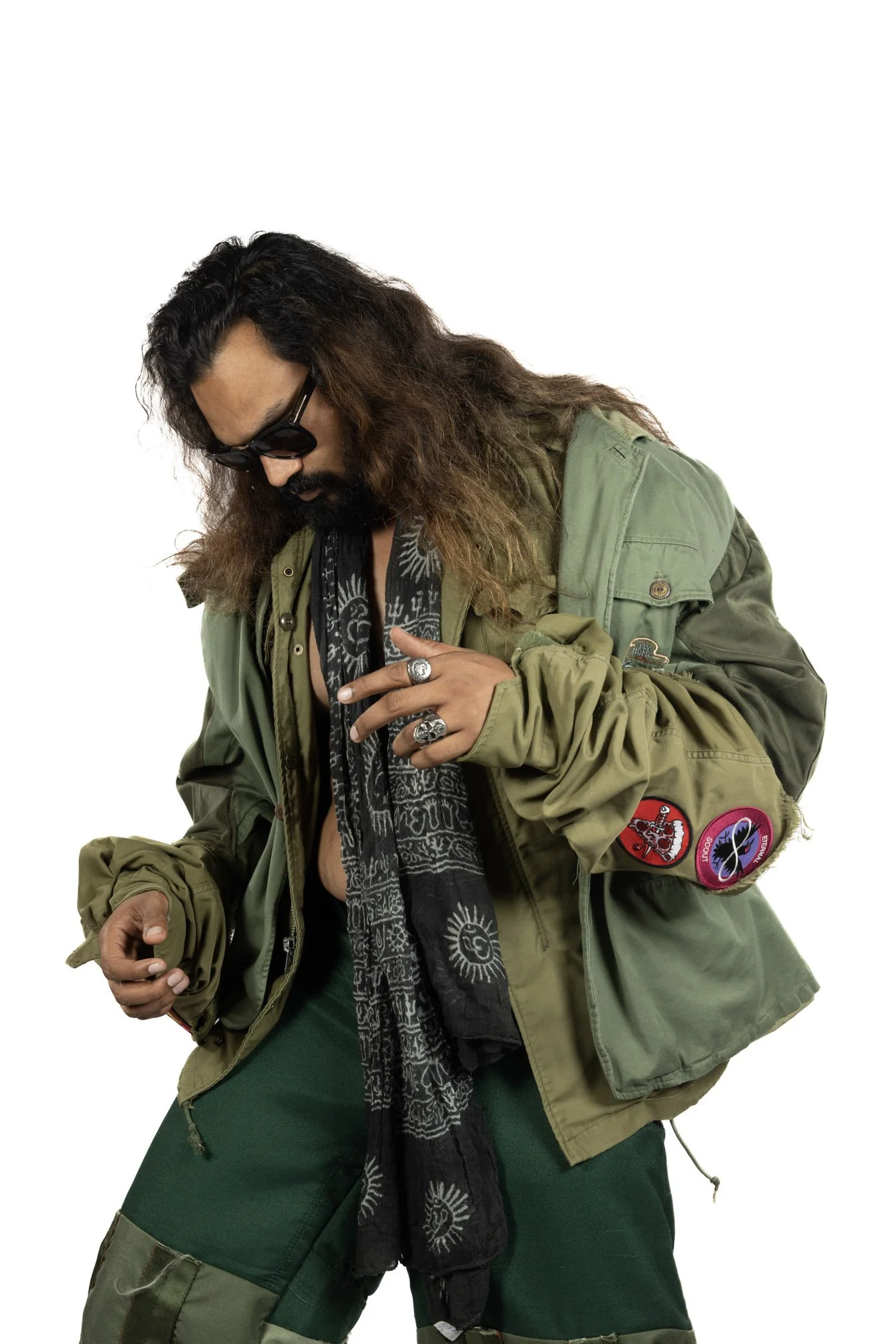 A man with long, wavy hair and sunglasses wearing a green military-style jacket, rings, and a black bandana.