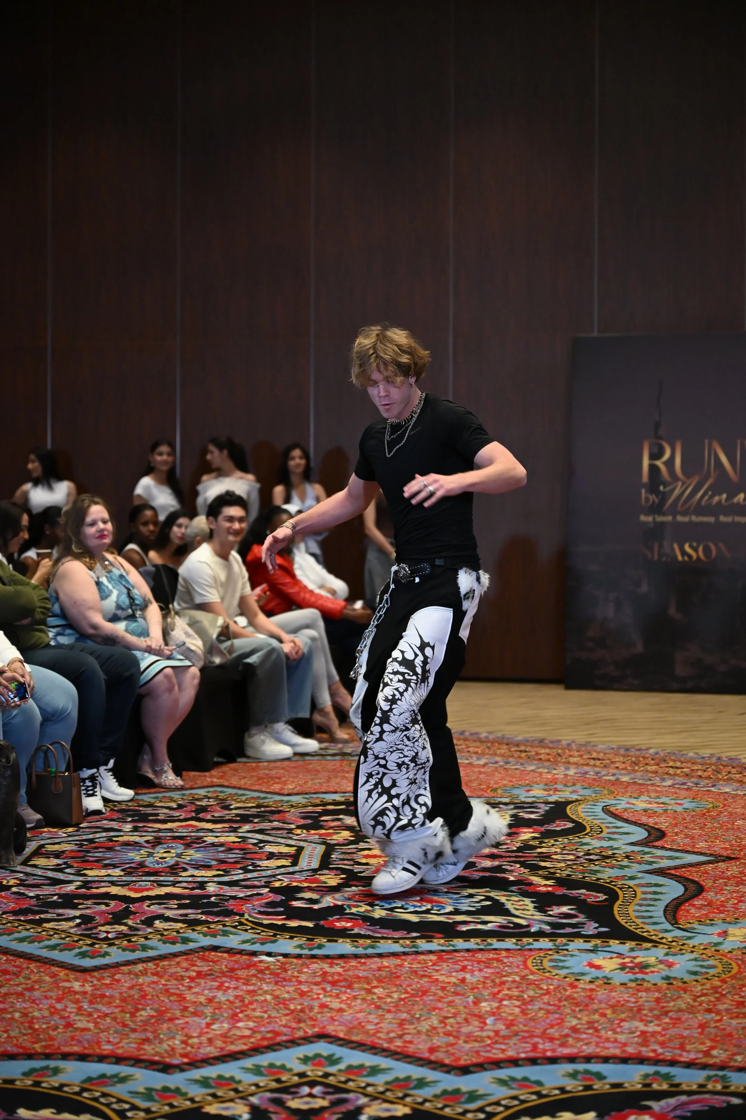 Person dancing on a patterned carpet at a fashion show with audience members watching.
