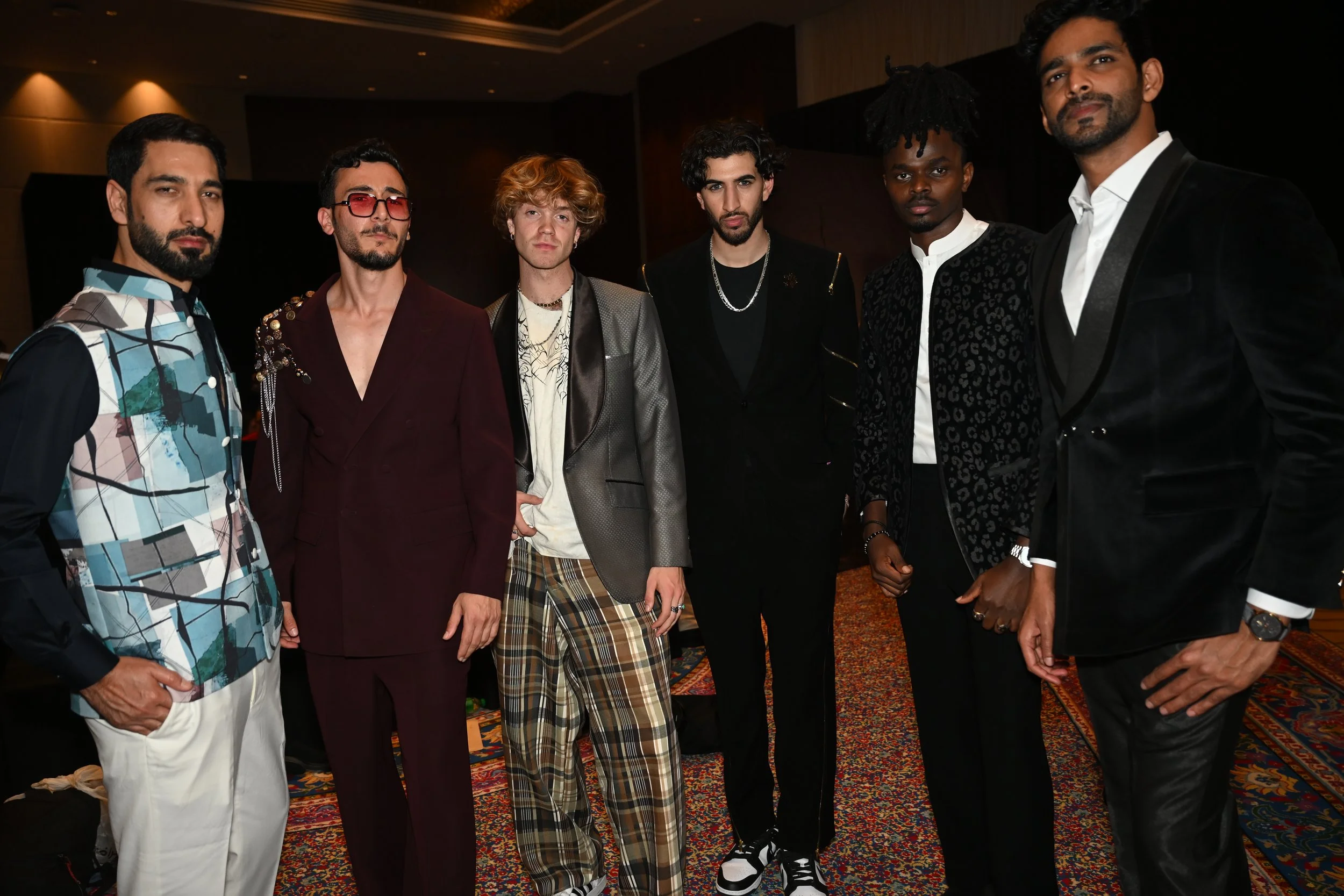 Group of seven men posing together in formal and stylish outfits at an indoor event.