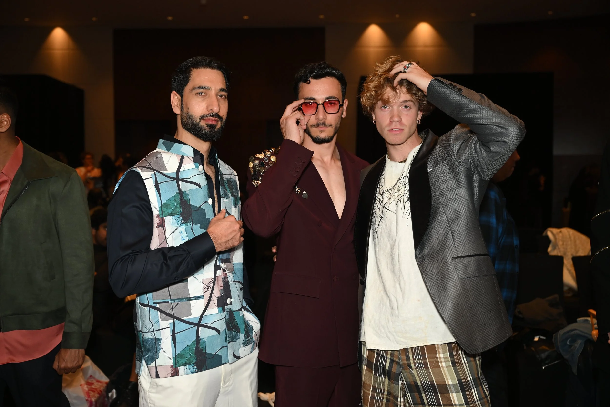 Three men standing together at an indoor event, dressed in stylish outfits, with one wearing a colorful patterned jacket, another in a maroon suit with sunglasses, and the third in a grey blazer with plaid shorts.