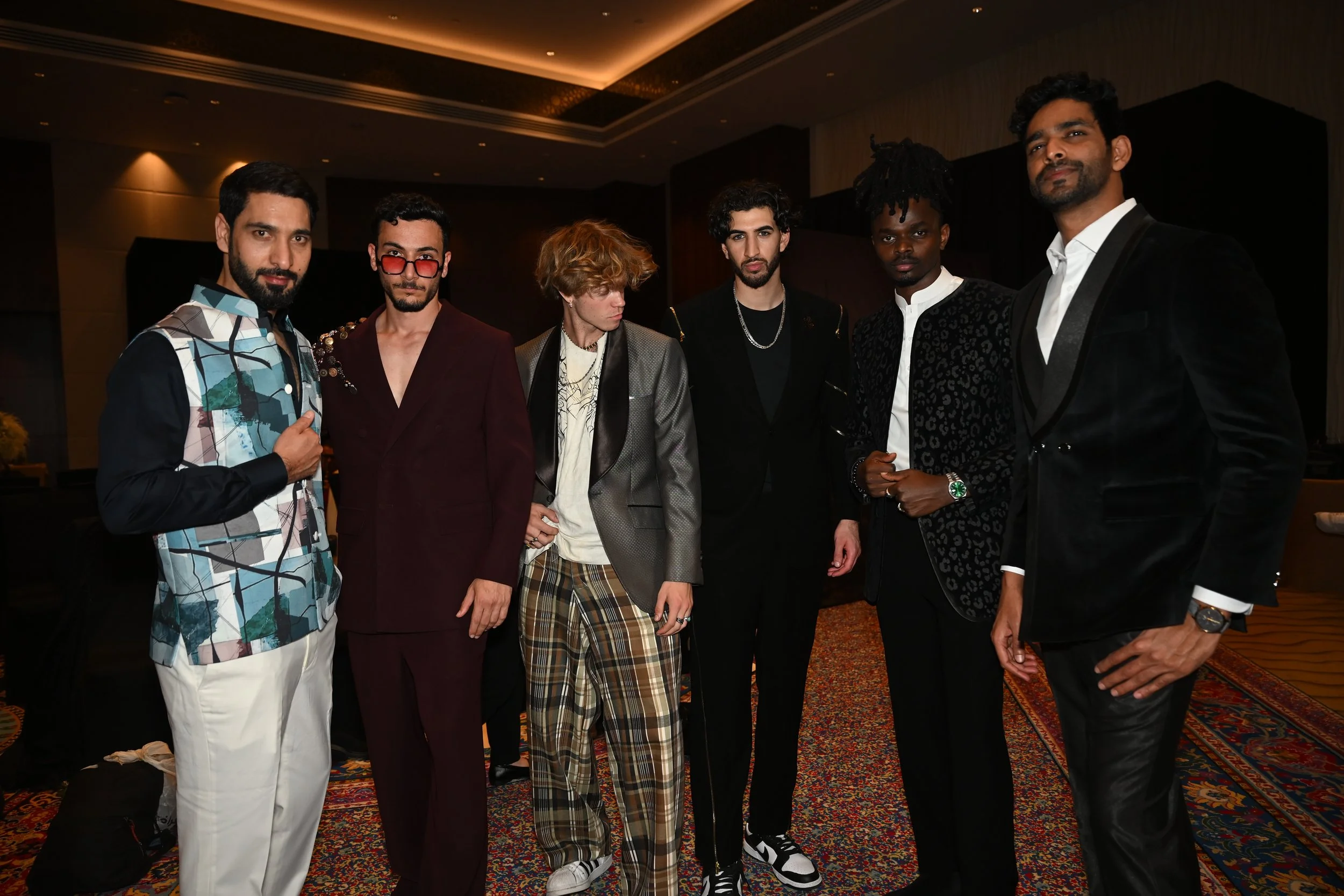Six men in fashionable clothing standing together in a dimly lit indoor setting, posing for a group photo.