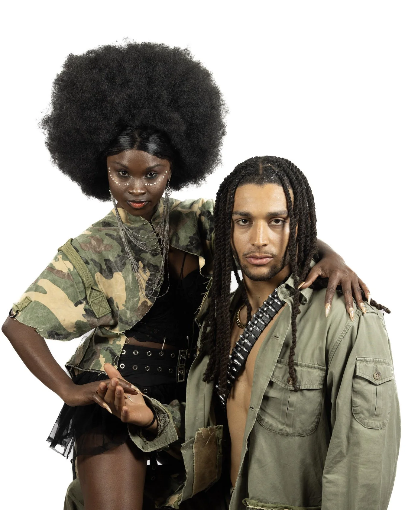 A woman with a large afro hairstyle and artful makeup wearing a camouflage jacket and jewelry, standing next to a man with long dreadlocks and an open green military-style jacket, on a white background.