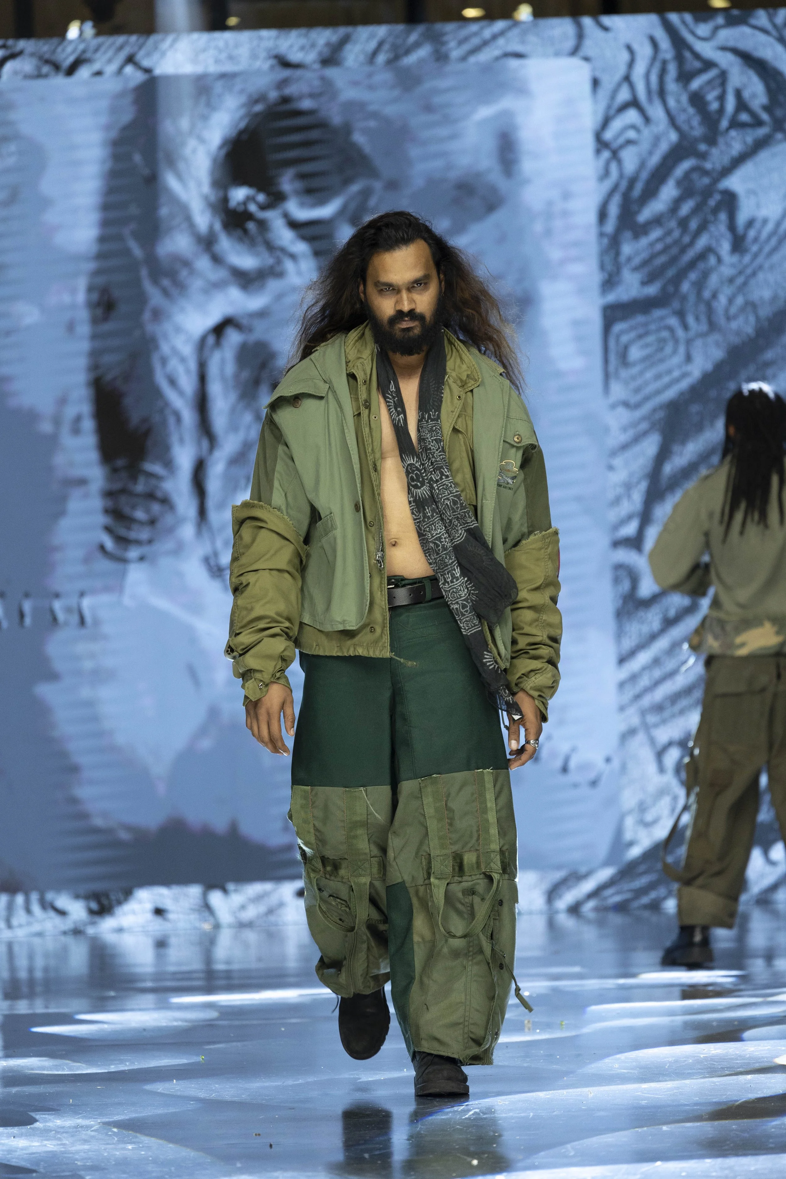 A male model with long hair and beard walking down a runway, dressed in military-style clothing, with a large digital screen displaying abstract black-and-white artwork in the background.