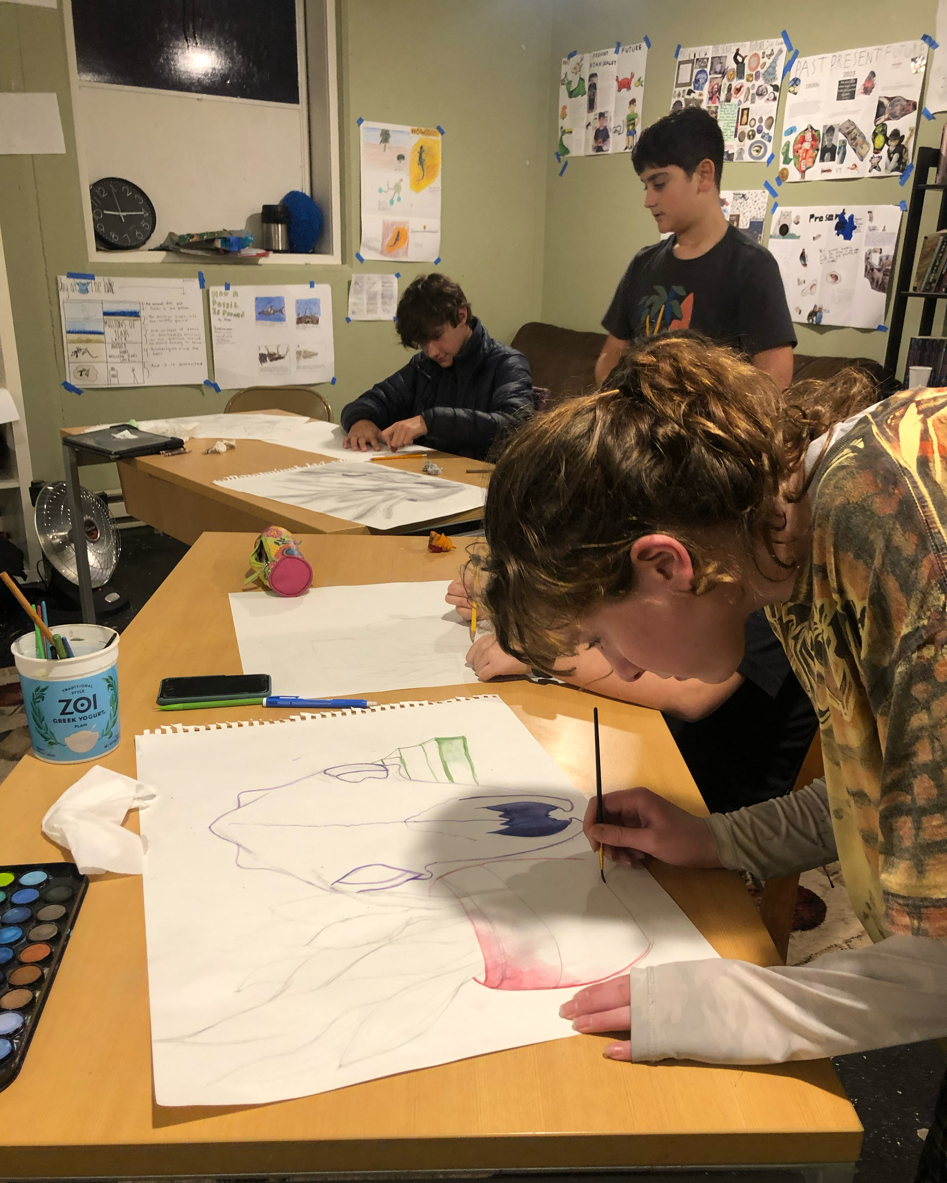 Multiple children and an adult are working on a large art project at a table, painting a big picture of a face.