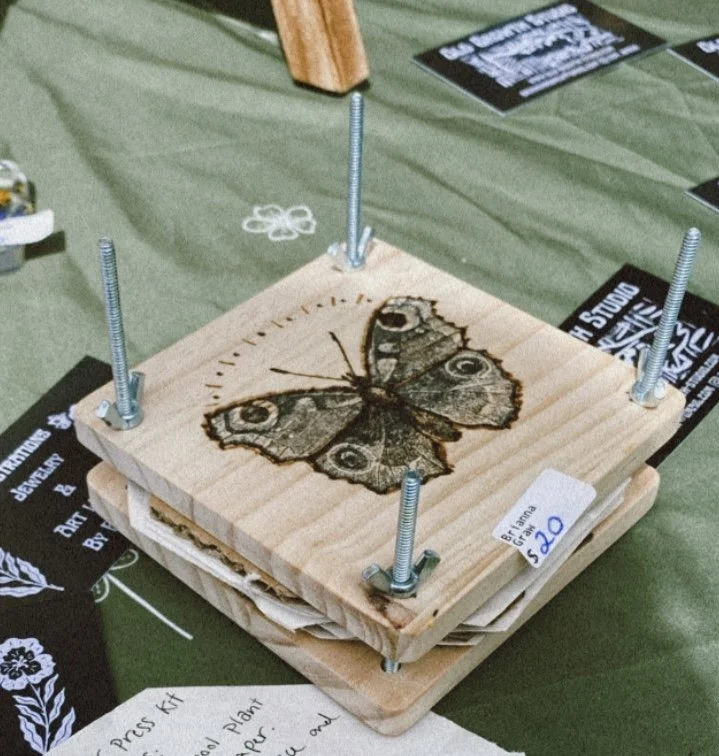 A wooden art piece with a butterfly design secured by four large bolts on a green tablecloth, with various informational and promotional papers surrounding it.