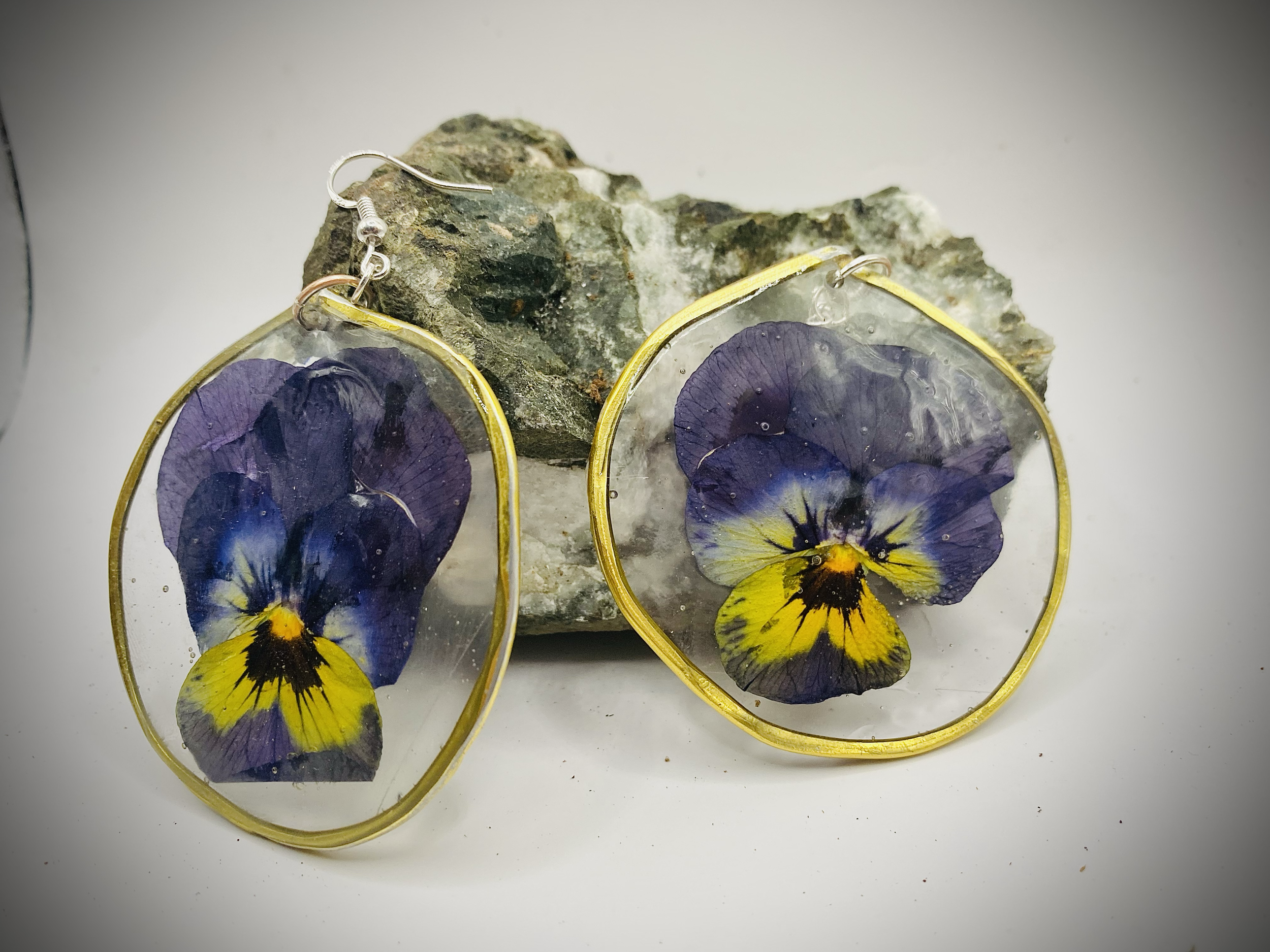 Earrings with pressed purple and yellow pansy flowers encased in clear resin and gold-colored metal, resting on a textured gray rock.