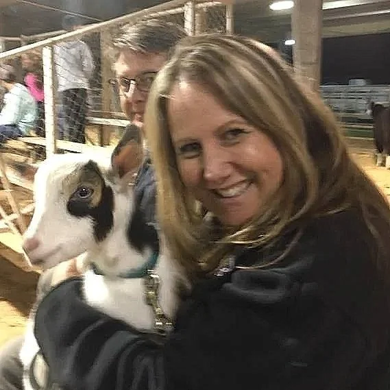 A woman holding a baby goat at a fair or farm, with a man and wooden bleachers in the background.