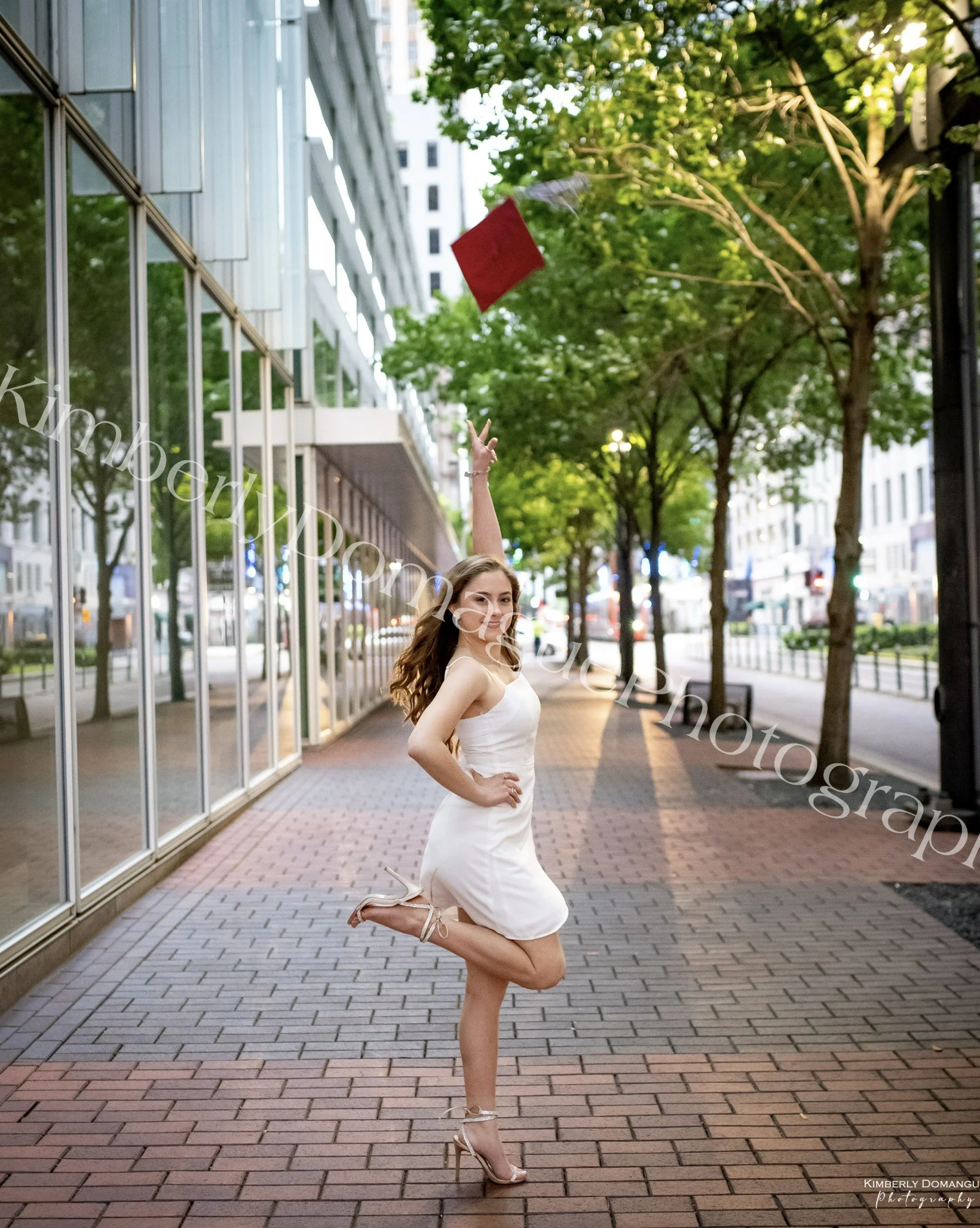 A woman dancing and throwing a red bag into the air on a city sidewalk lined with trees and modern buildings.