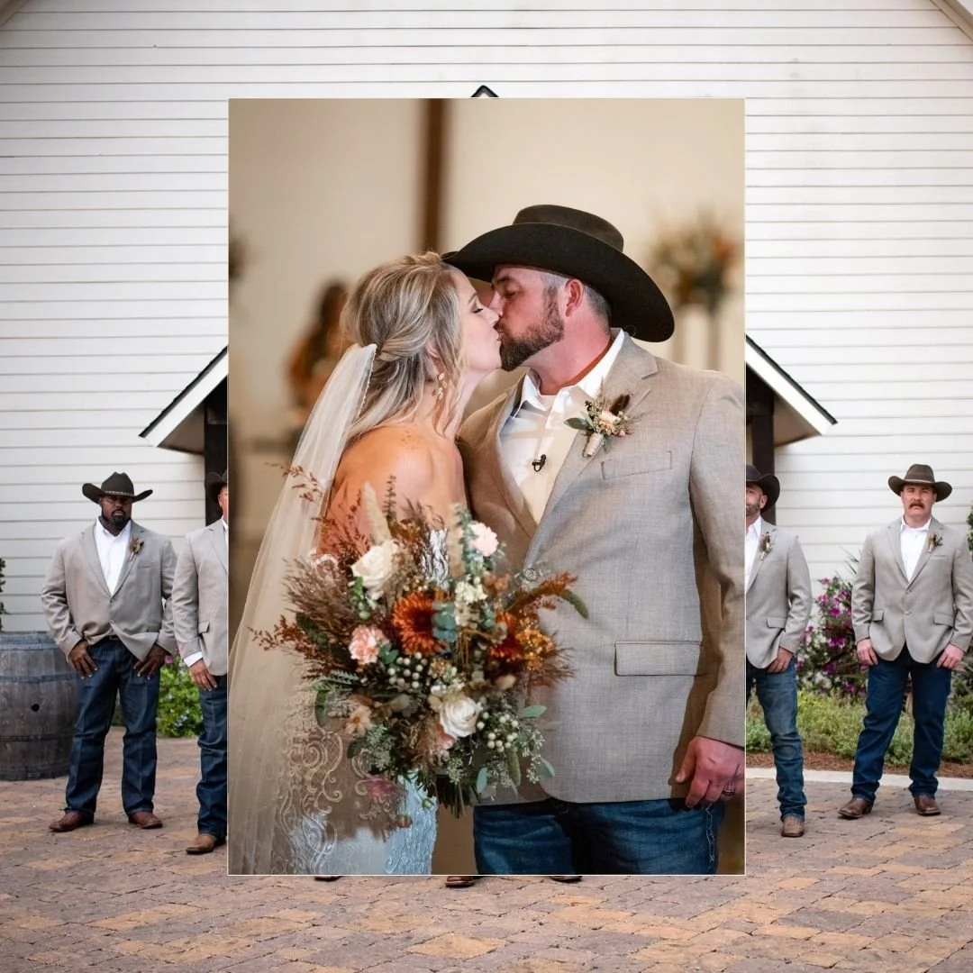 A wedding photo featuring a bride and groom kissing, with the bride holding a bouquet of flowers. The bride has blonde hair and is wearing a wedding dress with a veil. The groom has a beard and is wearing a gray suit, white shirt, and a black wide-br