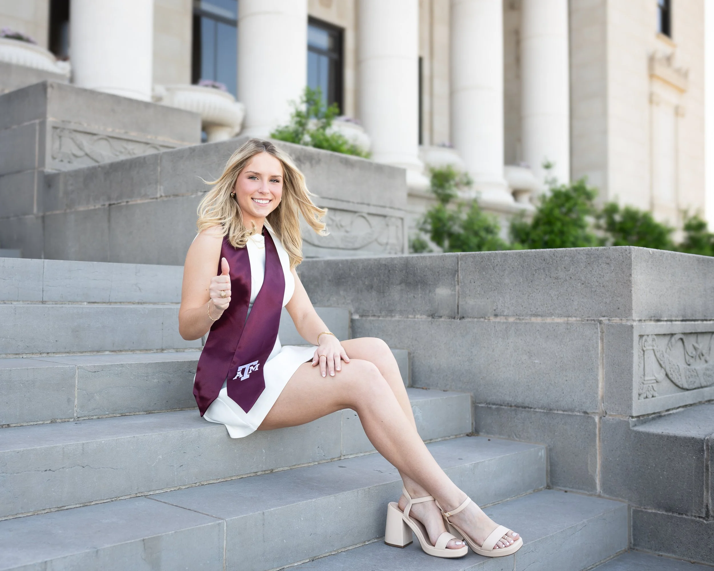 Young woman in a white dress with a maroon graduation stole sitting on steps outside a large official building, giving a thumbs-up and smiling.