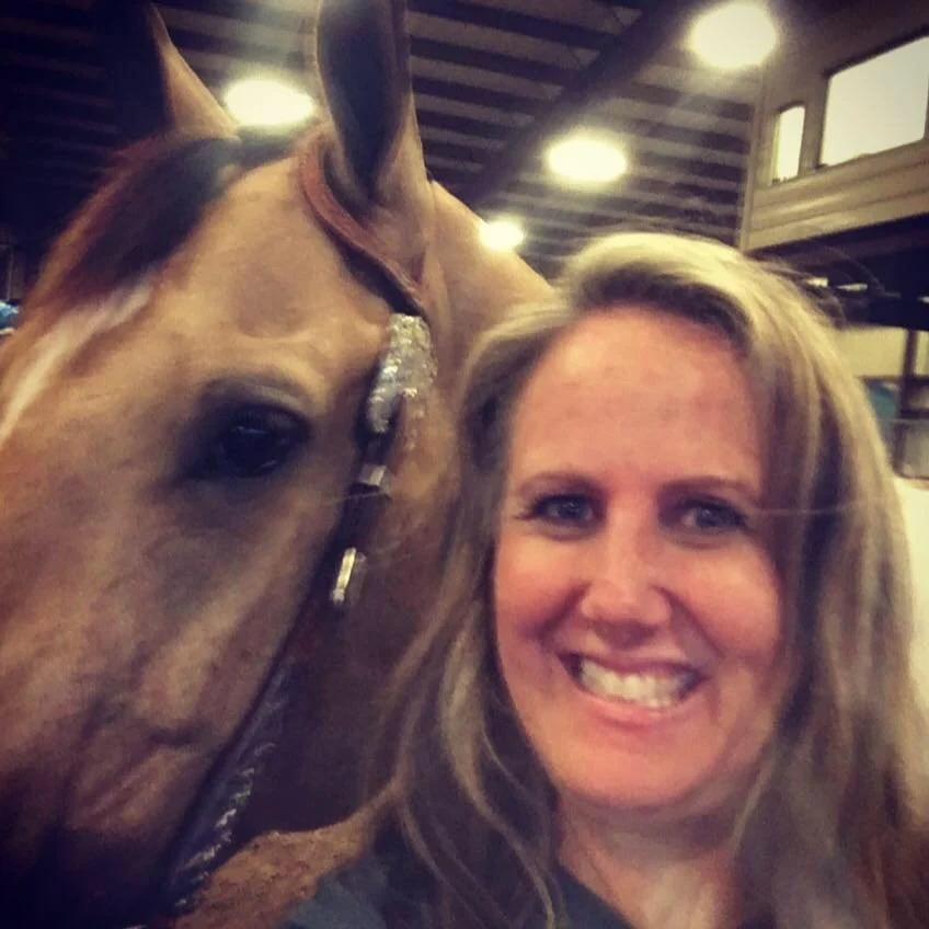 A woman smiling next to a horse in a barn or stable with wooden beams and indoor lights.