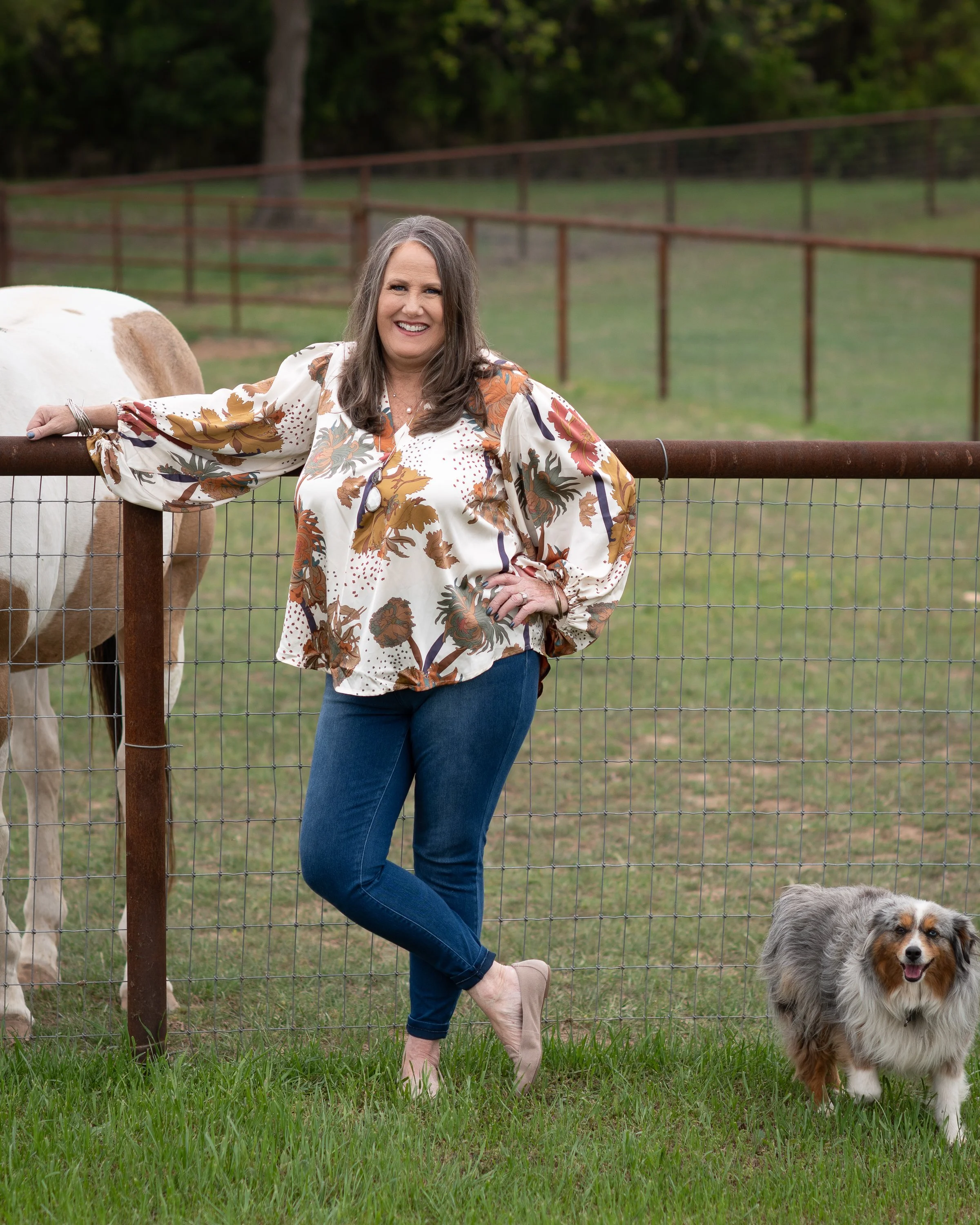 A woman with gray hair wearing a floral blouse and blue jeans, standing near a fence with a dog on a grassy field, with a cow behind the fence and trees in the background.