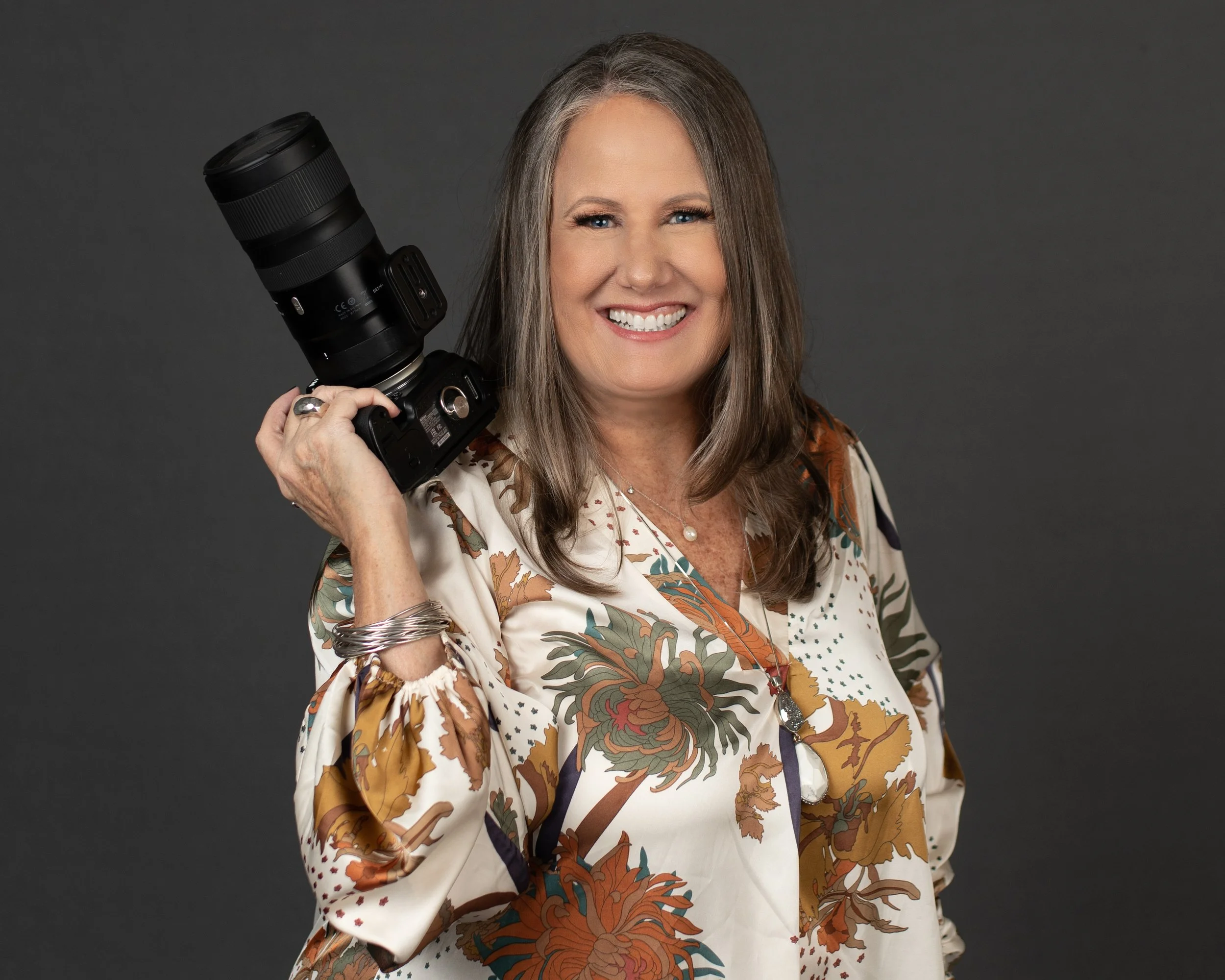A smiling woman with shoulder-length brown hair holding a professional camera with a large telephoto lens on her shoulder, against a dark gray backdrop.