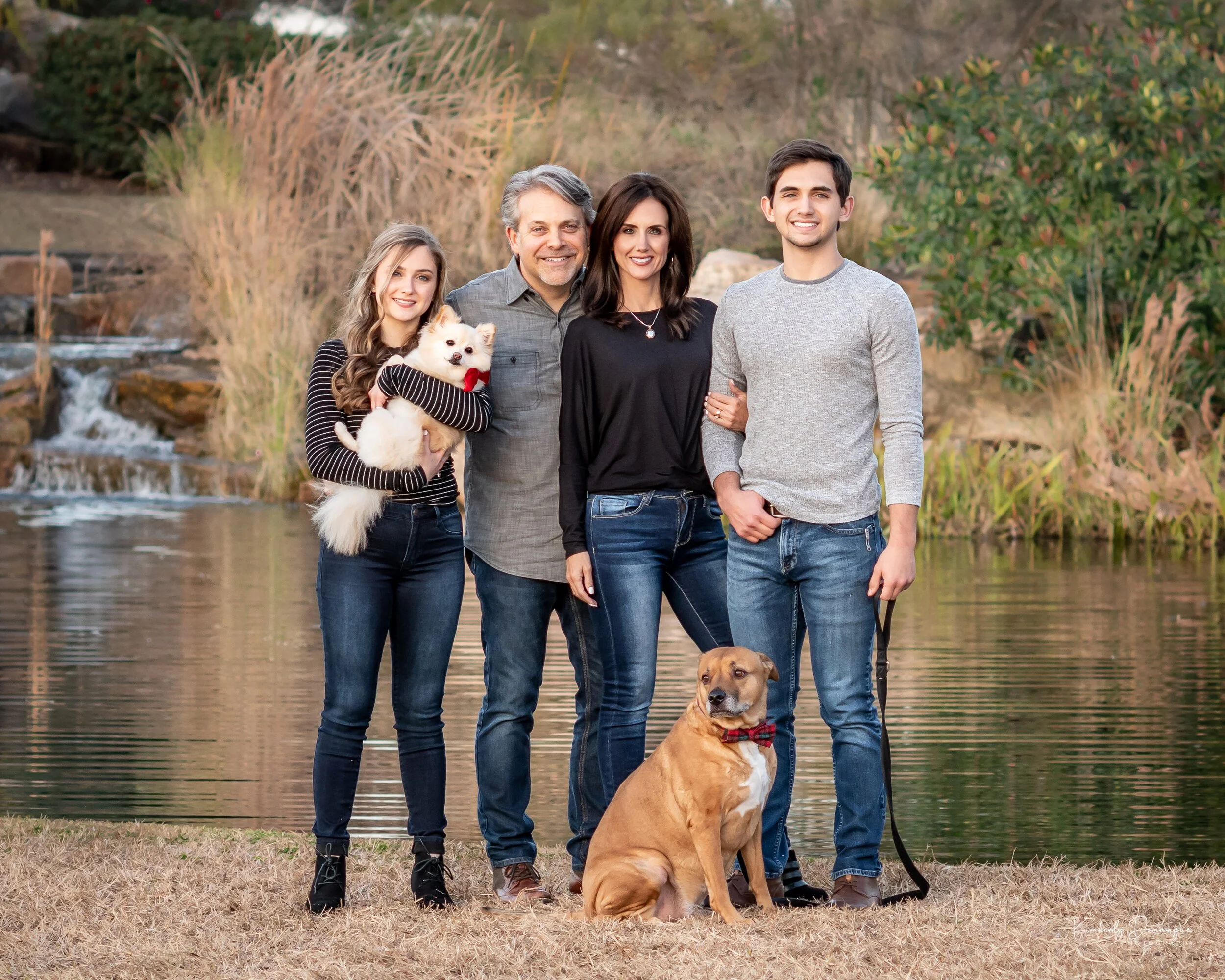 A family of five, including a dog and a puppy, posing outdoors near a river with trees and shrubs in the background.