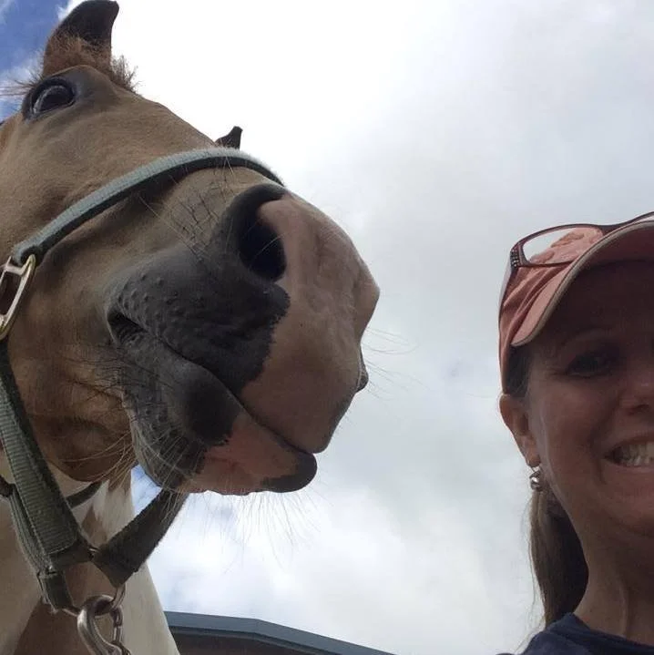 Close-up of a horse and a smiling woman in baseball cap posing outdoors on a cloudy day.