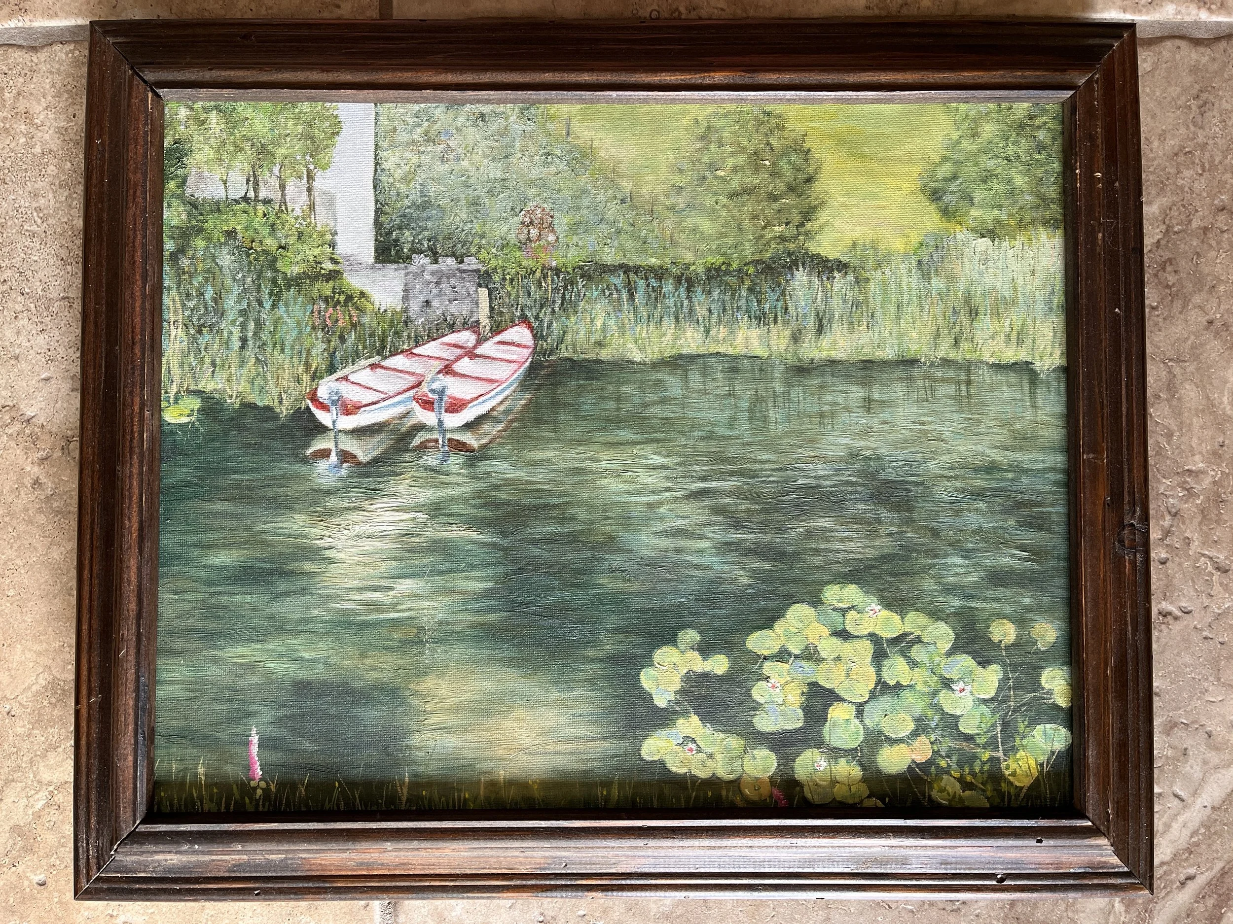 Painting of a lakeside scene with two paddle boats on water, surrounded by water lilies and trees, framed in dark wood.