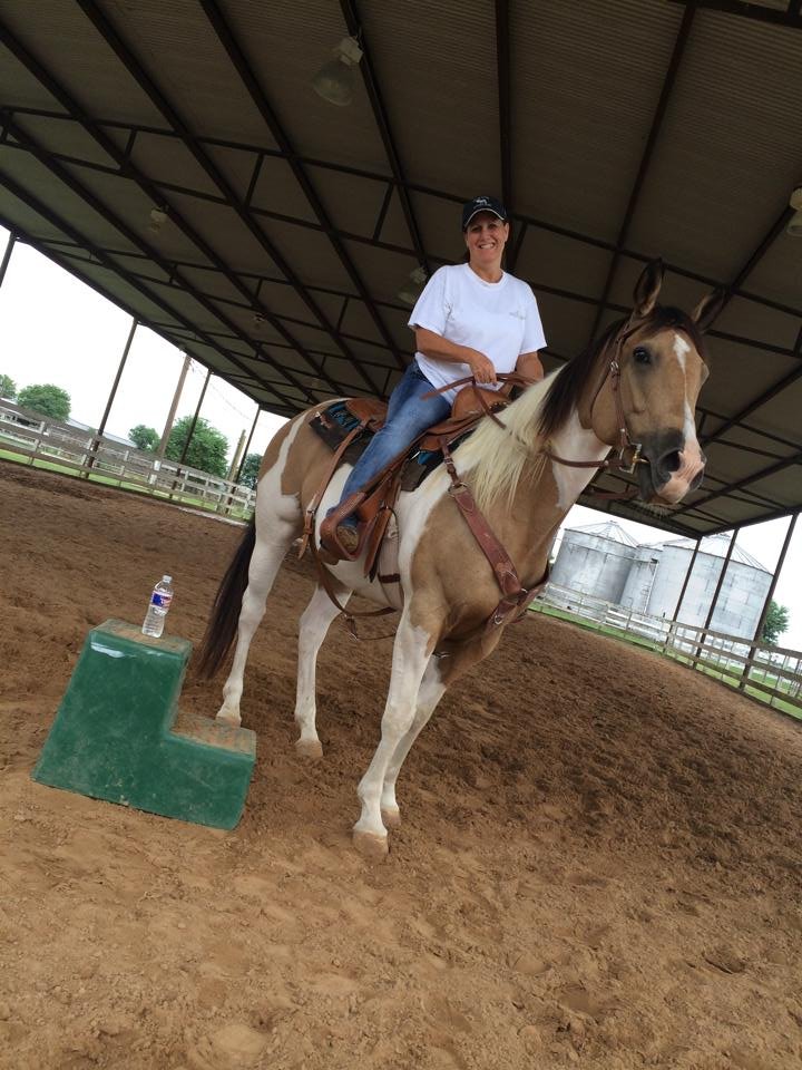 A woman smiling while sitting on a tan and white pinto horse with a brown saddle inside a large covered riding arena. There is a green step stool with a water bottle on top in front of the horse.