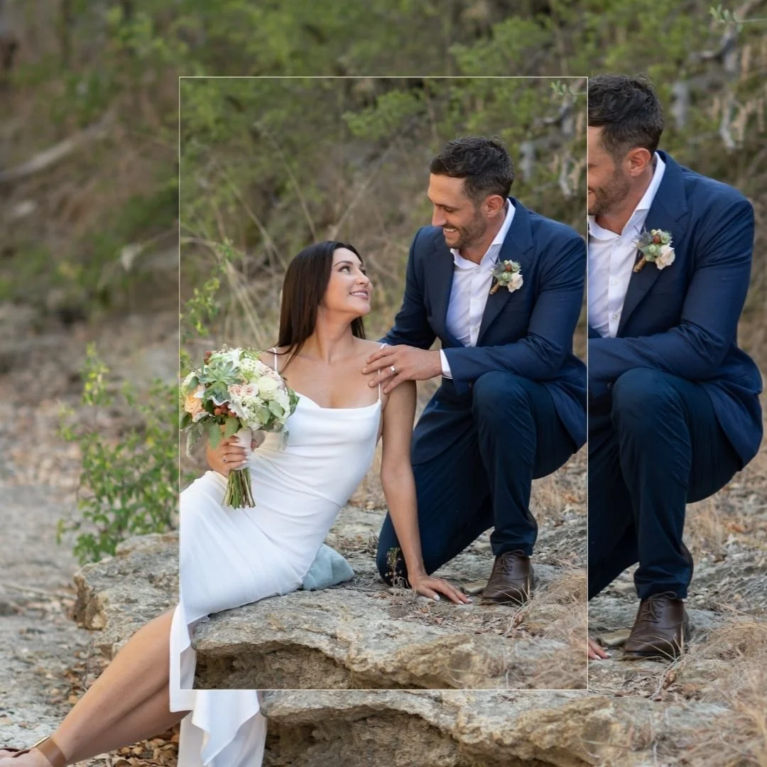 A bride and groom in wedding attire outdoors, with the bride holding a bouquet and smiling at the groom, who is crouching beside her.