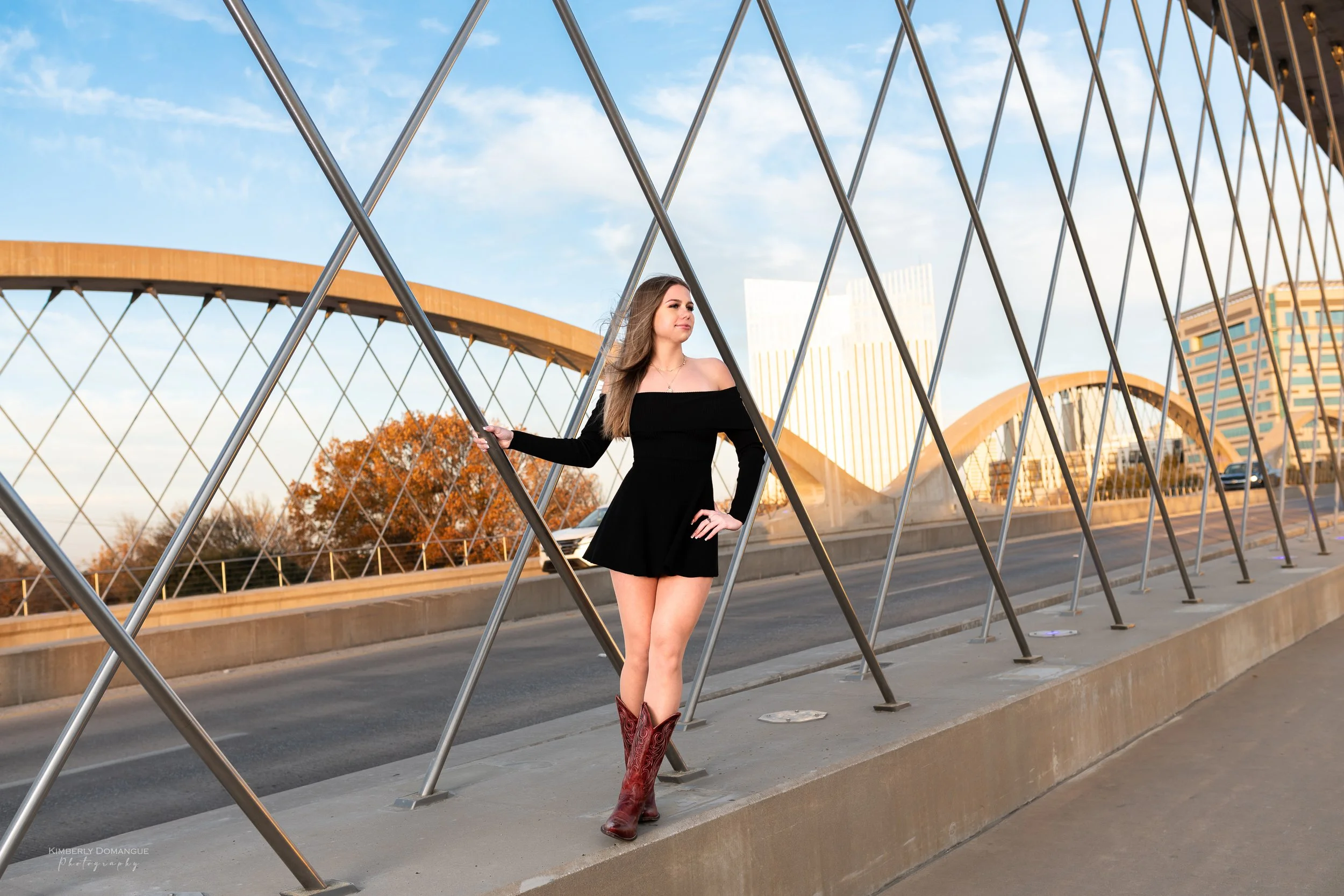 Woman in black dress and cowboy boots standing on a bridge with a modern, curved metal structure, city buildings in the background, and a blue sky with scattered clouds.
