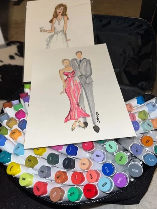 Two fashion illustrations on paper surrounded by colorful paint swatches, with a black carrying case in the background.