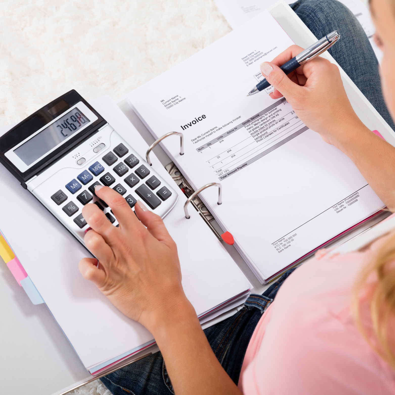 A person using a calculator and writing on an invoice with a pen, with folders and papers on a white surface.