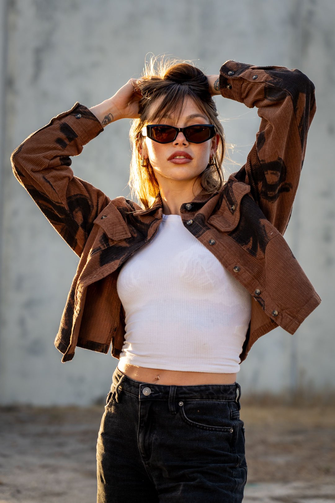 A young woman wearing black sunglasses, a white crop top, black jeans, and a brown camouflage-patterned jacket, standing outdoors against a gray concrete wall, with her hands in her hair.