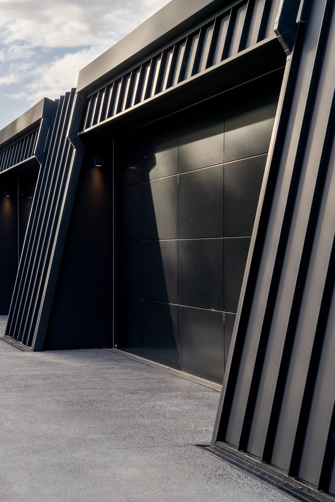 Modern building with dark metal paneling, vertical metal slats, and angled architectural design; sidewalk in foreground.