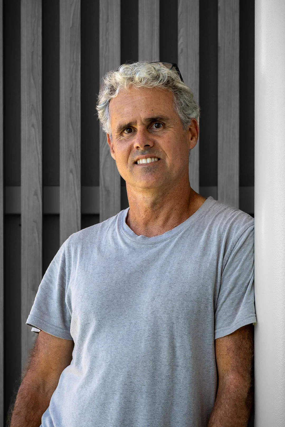 A middle-aged man with gray curly hair and glasses on his head, leaning against a white wall, wearing a light gray T-shirt, with a dark wooden slat background.