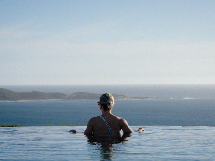 Person relaxing in an infinity pool overlooking the ocean with a distant shoreline and hills in the background.