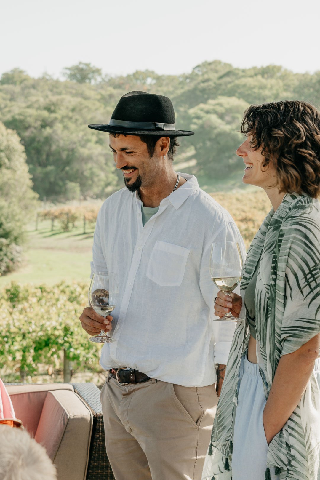 A man and woman smiling and holding glasses of wine outdoors with trees and a grassy landscape in the background.