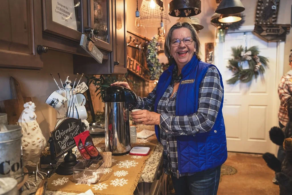 A woman with glasses and gray hair, smiling, stands behind a coffee station at a festive holiday event, wearing a blue vest and plaid shirt.