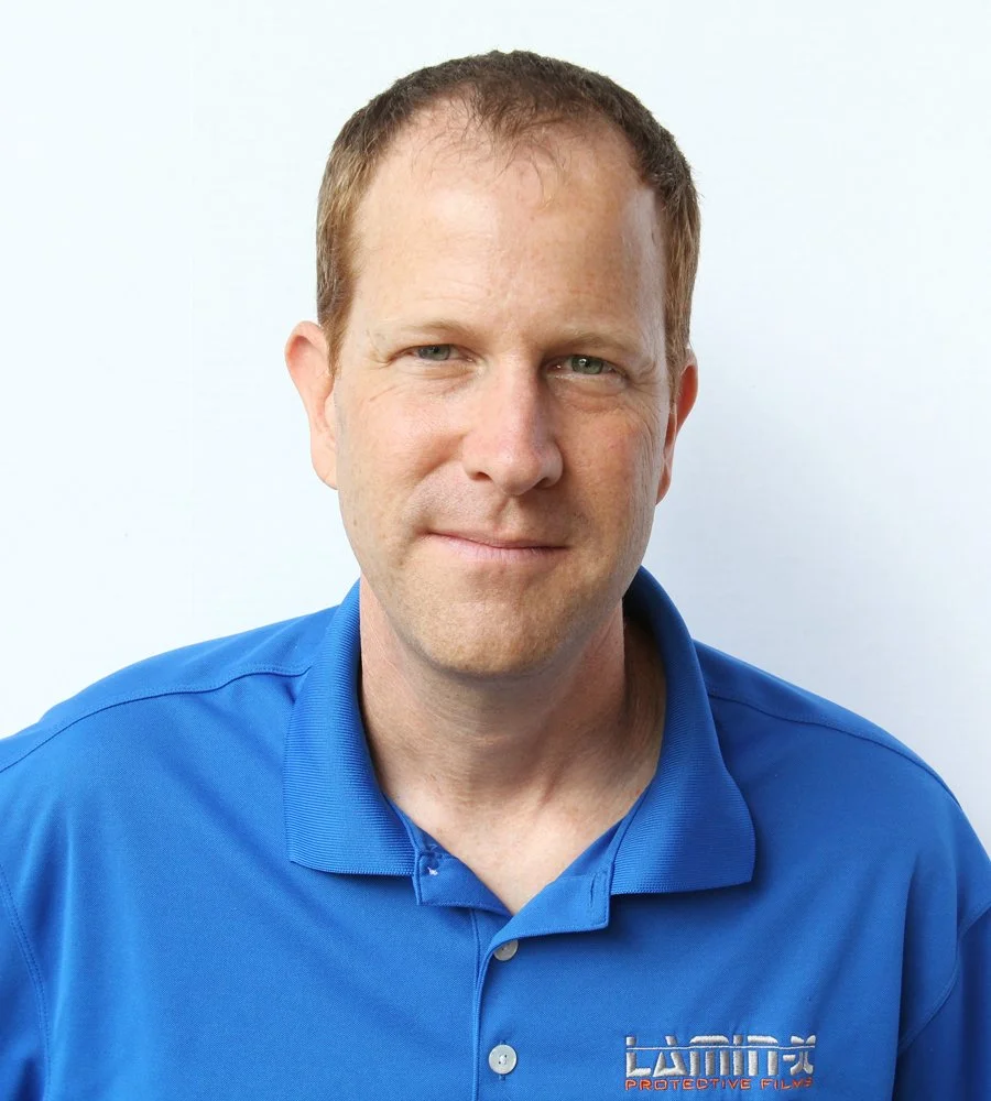 Portrait of a man with short light brown hair wearing a blue collared polo shirt with a logo, smiling slightly against a plain white background.