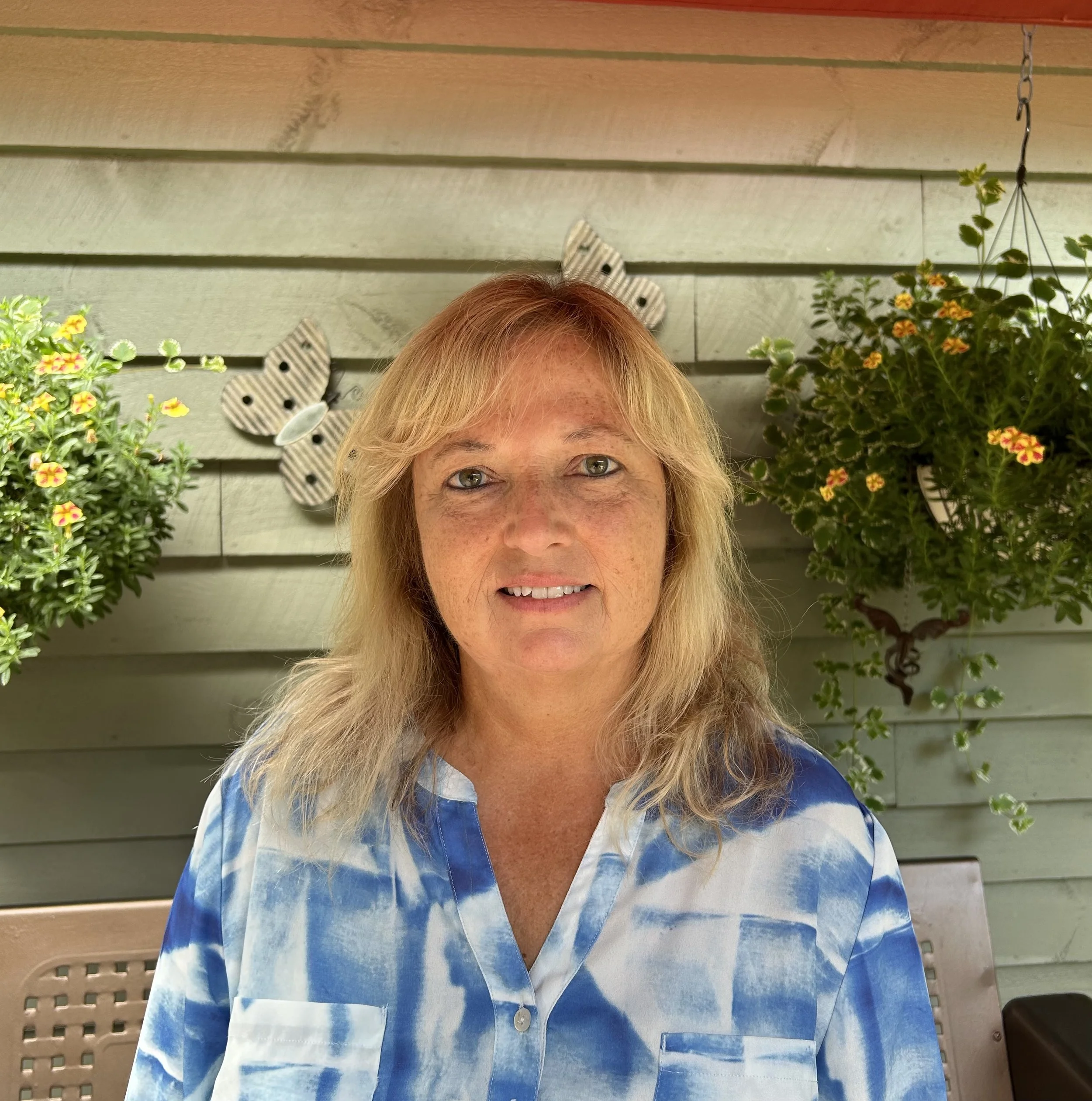 A woman with blonde hair and fair skin sitting outdoors in front of a green wooden wall, flanked by hanging and potted flowering plants, with butterfly decorations on the wall behind her.