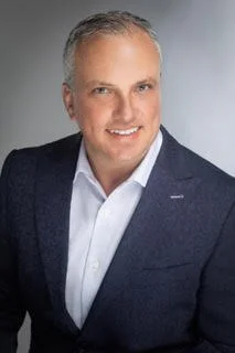 Portrait of a smiling middle-aged man with short gray hair, wearing a dark blazer over a white shirt against a gray background.