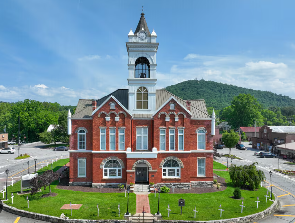 A historic red brick courthouse with a clock tower, surrounded by a grassy lawn with white crosses, in a small town setting with mountains in the background.