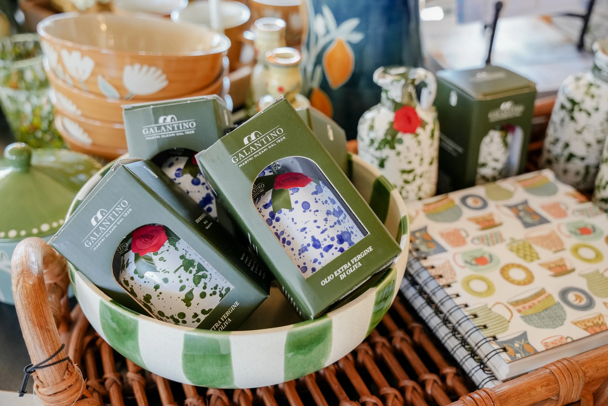 A display of various kitchen items, including boxed bottles of extra virgin olive oil with green and white splatter designs, ceramic bowls with floral patterns, and a patterned notebook on a woven basket.
