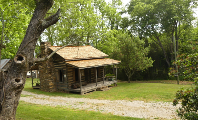 A rustic cabin with a small porch, situated in a lush green yard with trees and a gravel driveway.