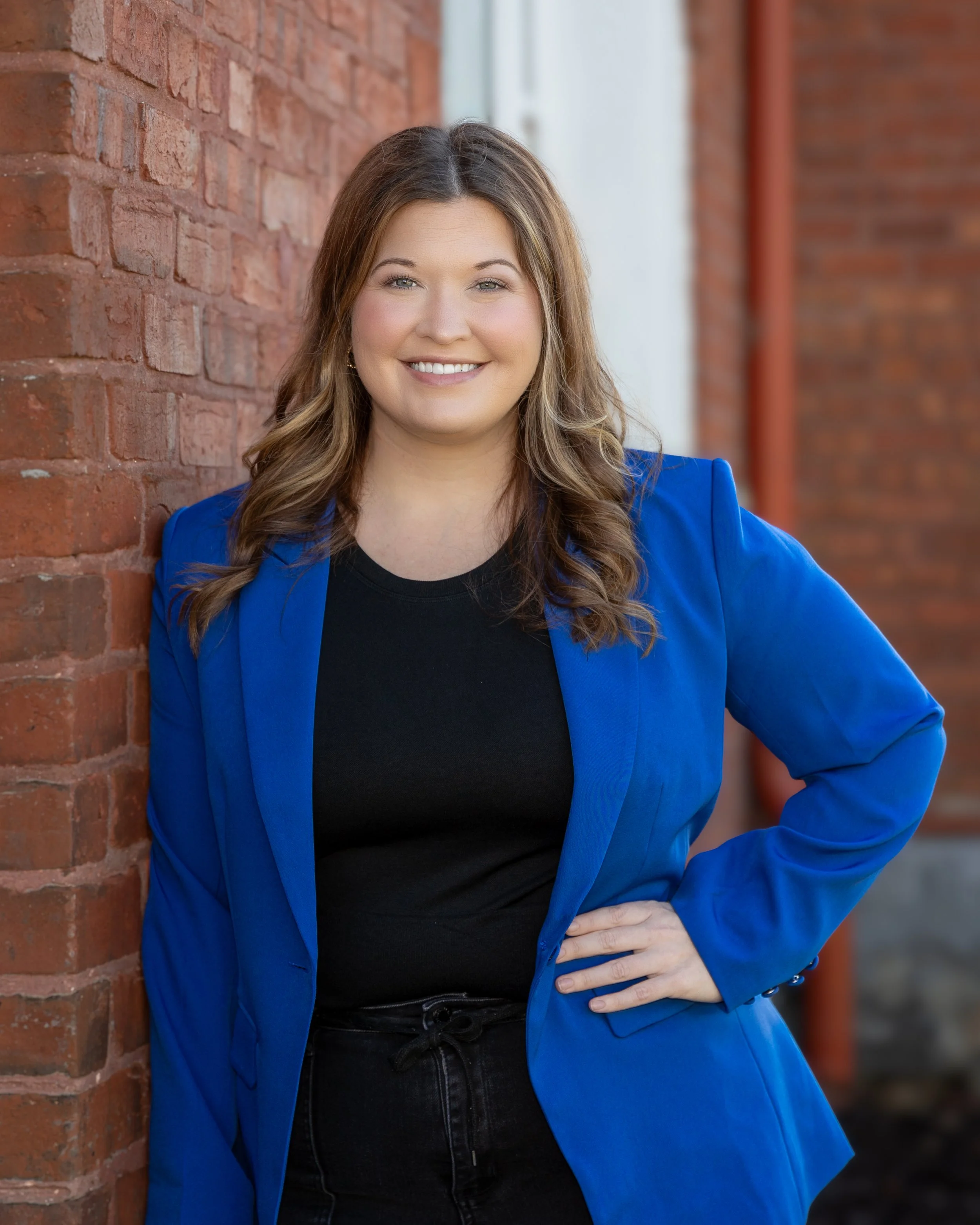 A woman with long wavy brown hair smiling, wearing a bright blue blazer over a black top, standing outdoors next to a brick wall.
