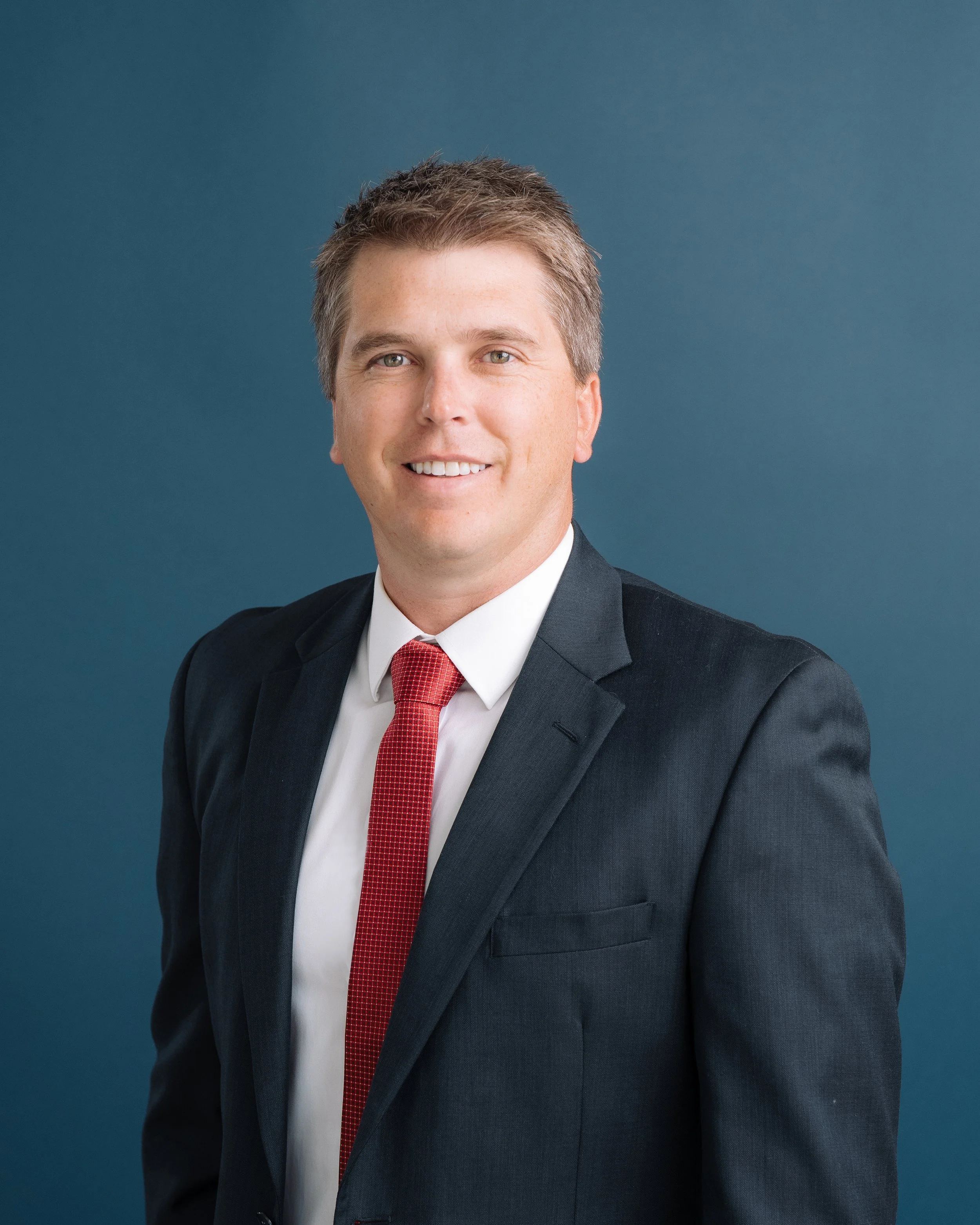 A man in a dark suit and red tie smiling, against a blue background.