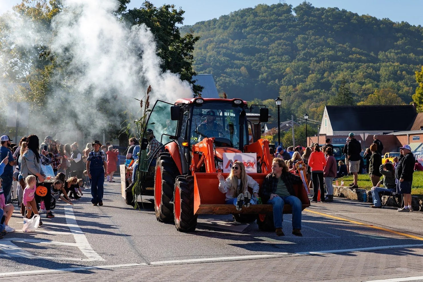 People enjoying a parade with a large orange tractor on the street, surrounded by spectators, some dressed in Halloween costumes, and mountains and buildings in the background.