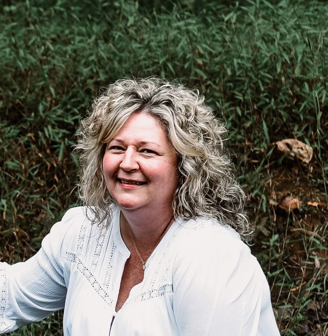 Smiling woman with long dark hair, wearing a white sweater, maroon cardigan, and black leather pants, standing against a plain white wall.