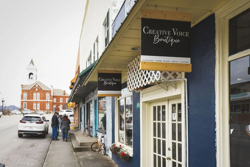 Street view of a boutique named 'Creative Voice Boutique' with signs for clothing, home goods, and music lessons. Pedestrians and parked cars are visible, with a red brick building and a water tower in the background.
