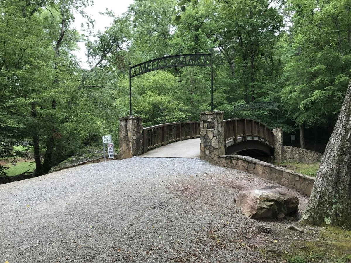 A small bridge with a stone and wood railing spans a small creek in a lush green forest. A sign reading "Bruce Bridge" is visible near the entrance.