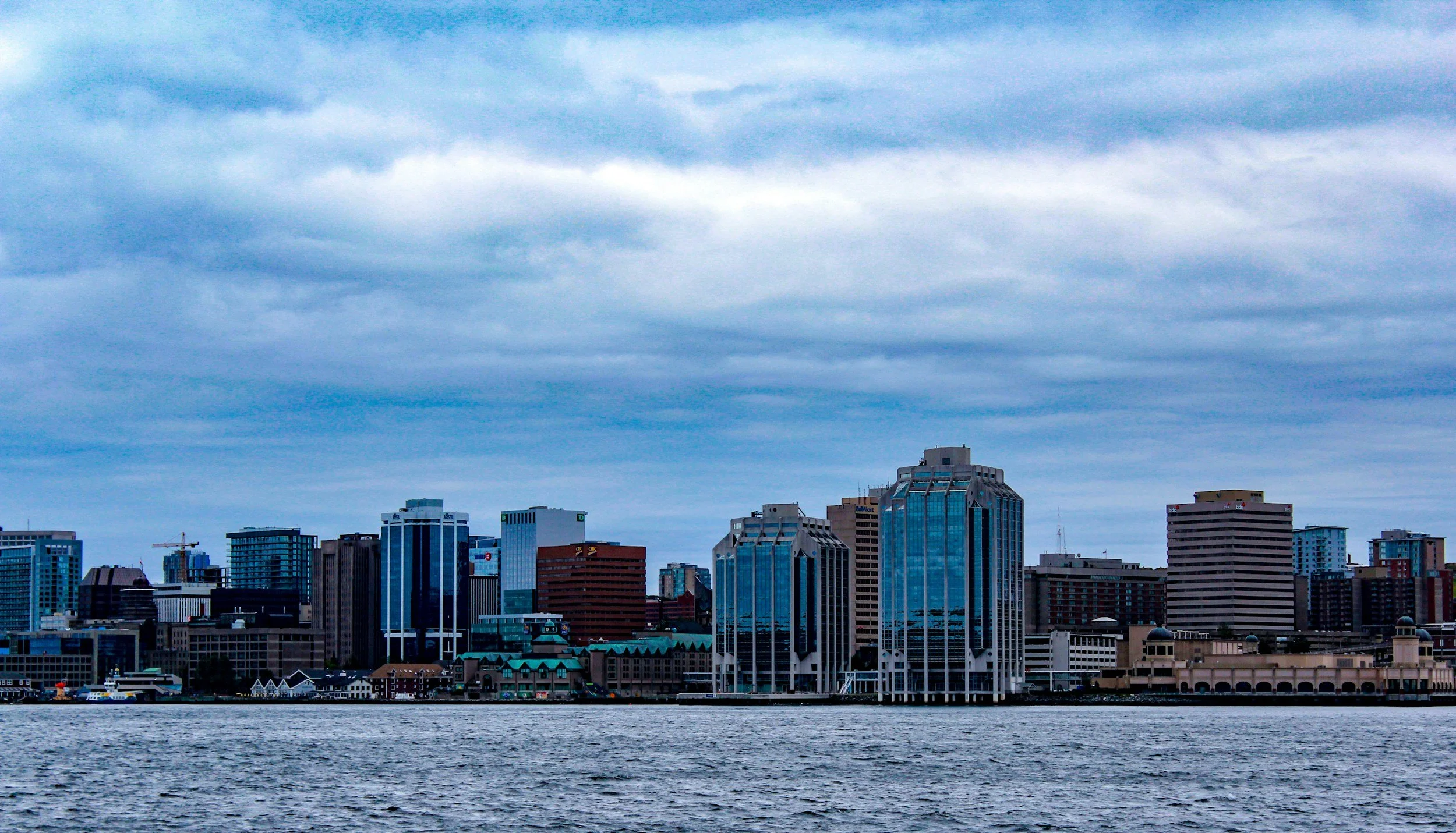 City skyline with various high-rise buildings along a body of water under cloudy sky.