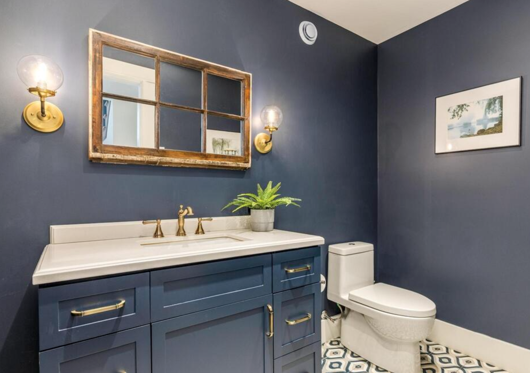 A modern bathroom with navy blue walls, a white vanity with gold handles and fixtures, a potted green plant, a white toilet, a decorative framed mirror, wall sconces with bulbs, and a landscape painting on the wall.