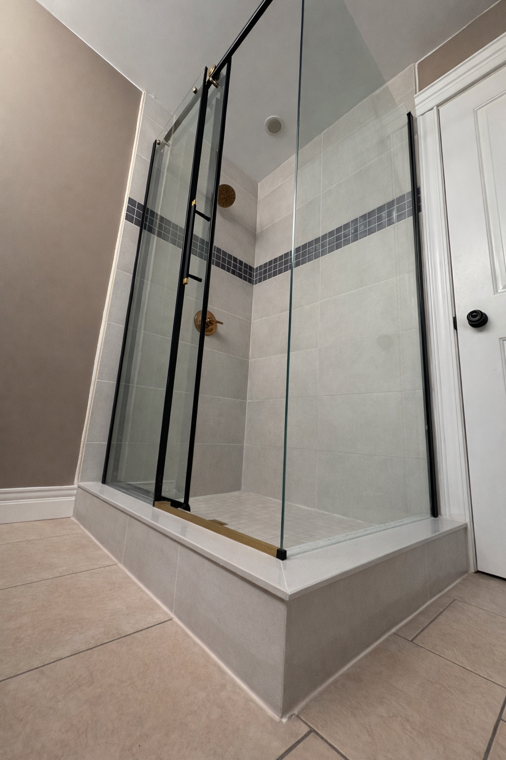 A modern glass shower enclosure with a black frame and a bronze showerhead inside a bathroom with beige tile walls and tan floor tiles. Tiled shower Halifax, Dartmouth, Bedford, Lower Sackville, Hammonds Plains, Fall River, Timberlea