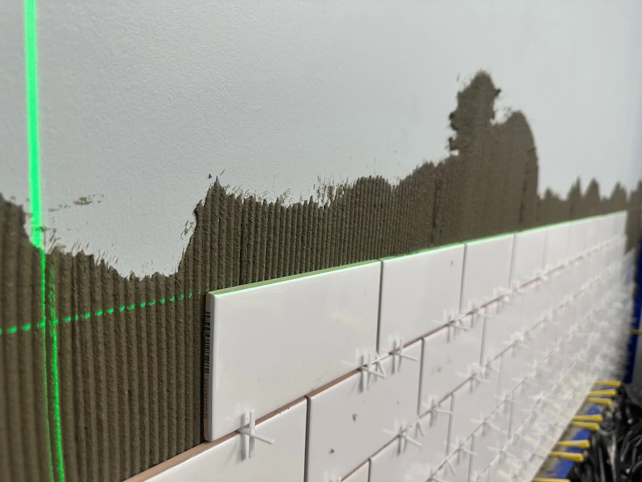 Close-up view of wall tiles and drywall installation, showing a green laser level and partially completed tile wall with spacers in place.