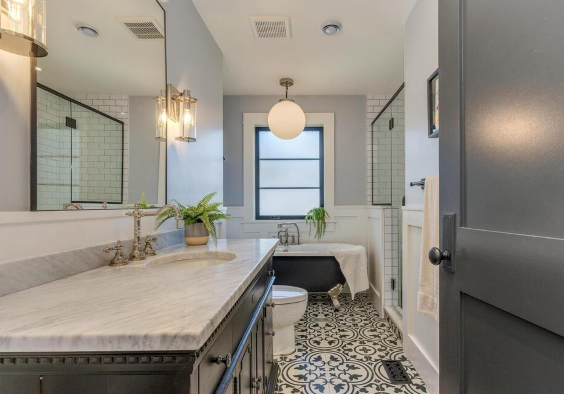 Modern bathroom with gray walls, black and white patterned floor, marble vanity with gold fixtures, large mirror, and a bathtub by the window with plants, and a shower area with glass door.