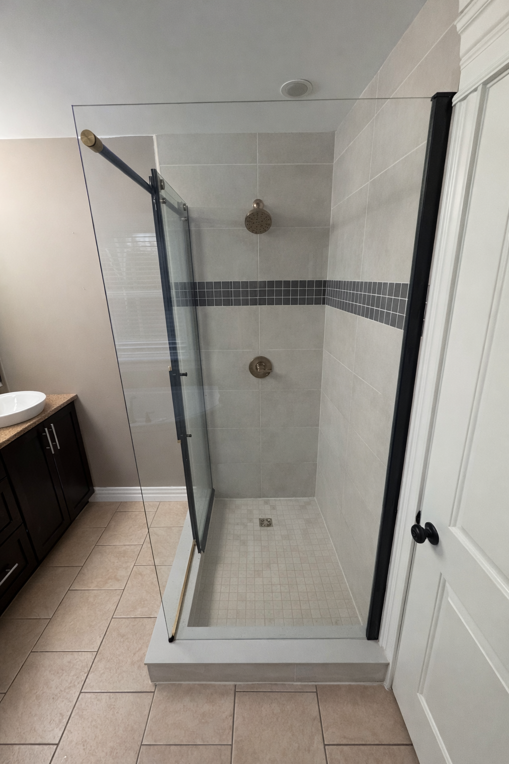 Shower with glass enclosure in a bathroom, with shower head and control handle, beige tile walls with a horizontal gray tile accent, and beige floor tiles. Shower Installation Halifax, Dartmouth, Bedford, Lower Sackville, Hammonds Plains, Fall River,