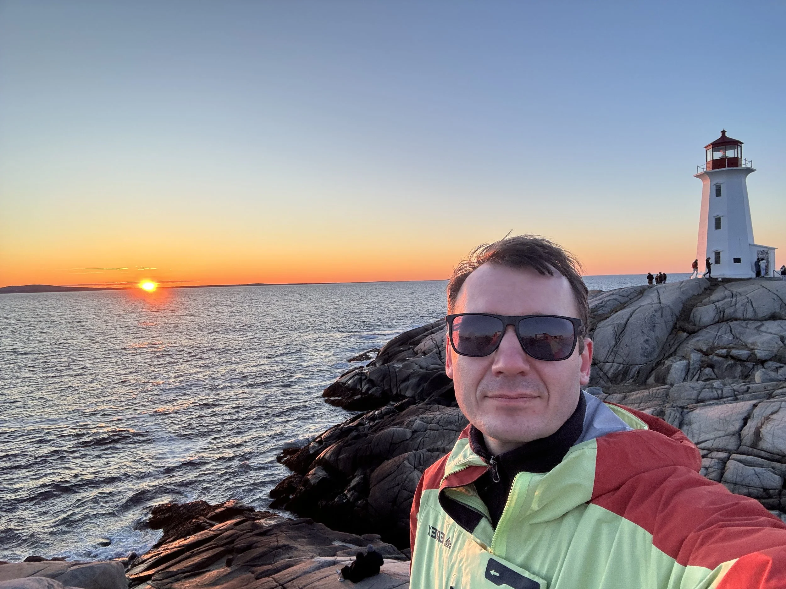 A man wearing sunglasses and a neon yellow and red jacket takes a selfie near a rocky coastline during sunset, with a lighthouse in the background and a few people standing on the rocks.